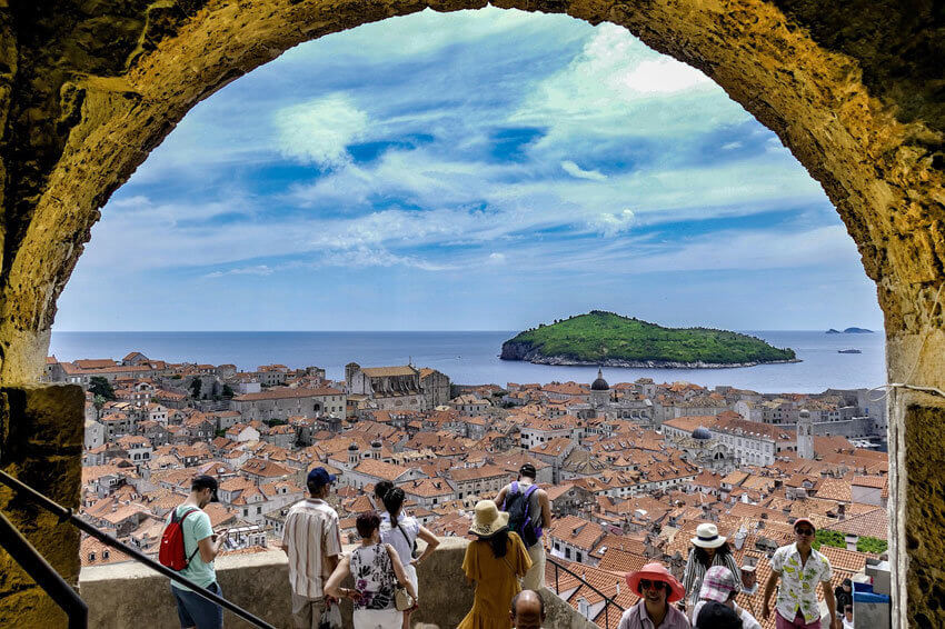 Blick durch einen steinernen Torbogen auf die Stadt und einen Felsen im Meer. Rouristen sind von weitem zu sehen